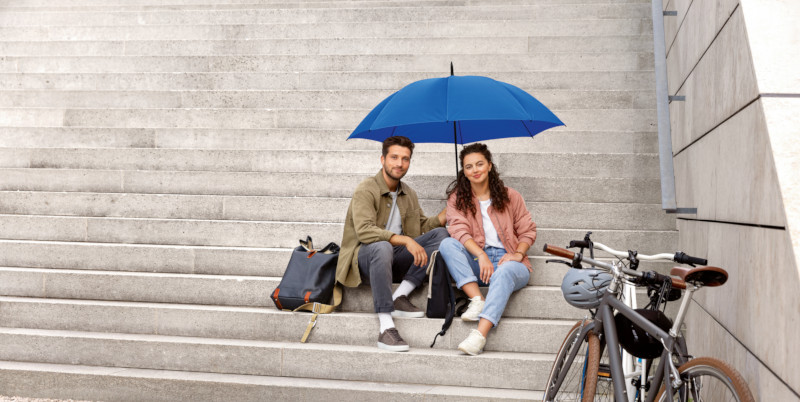Zwei Personen auf einer Treppe sitzend mit einem blauen Schirm über ihnen und einem Fahrrad vor ihnen