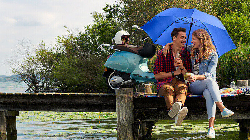 Zwei Personen auf einem Steg unter einem blauen Schirm mit einem Moped im Hintergrund